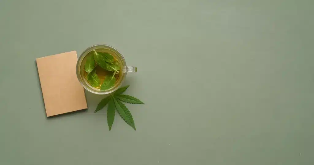 Cannabis leaf beside a cup of tea and a notebook on a table